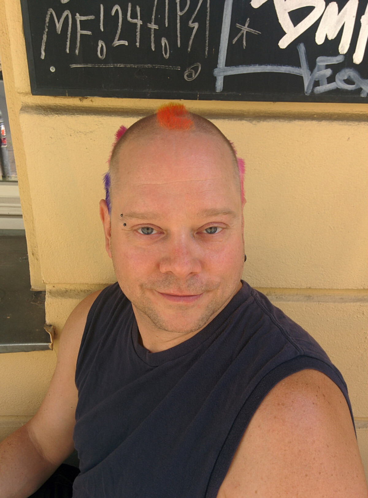 Rob smiling at the camera at a cafe, mostly shaved head with small colorful tufts: orange-red at the forehead center, pink patches above the temples, purple near the right temple. Bridge piercing, sleeveless dark t-shirt. Chalkboard in background.