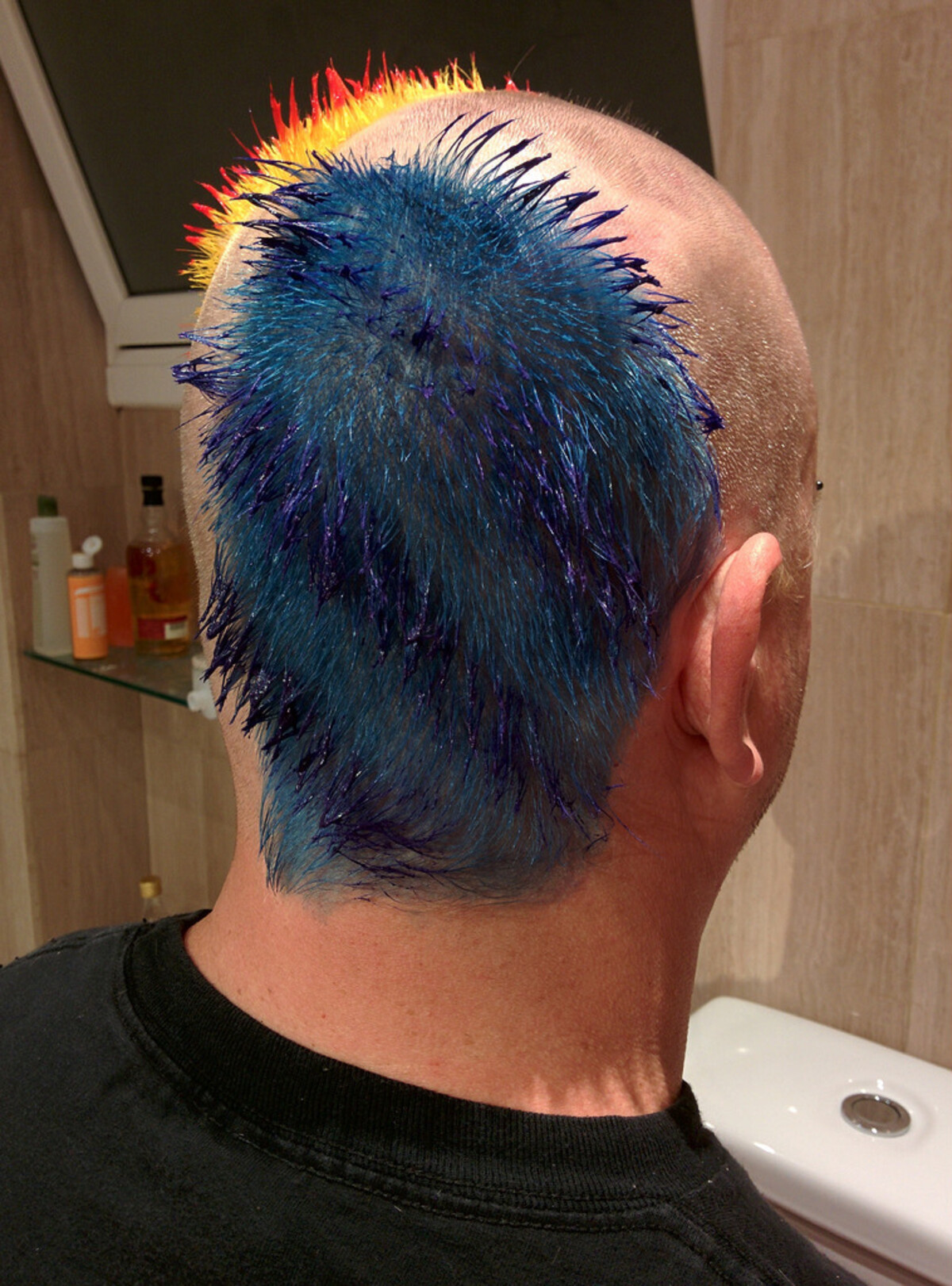 Back of Rob's head showing a mohawk strip: deep teal-blue spiky hair covering the back, transitioning sharply to bright orange and red at the crown. Sides fully shaved. Bathroom shelf with products visible.