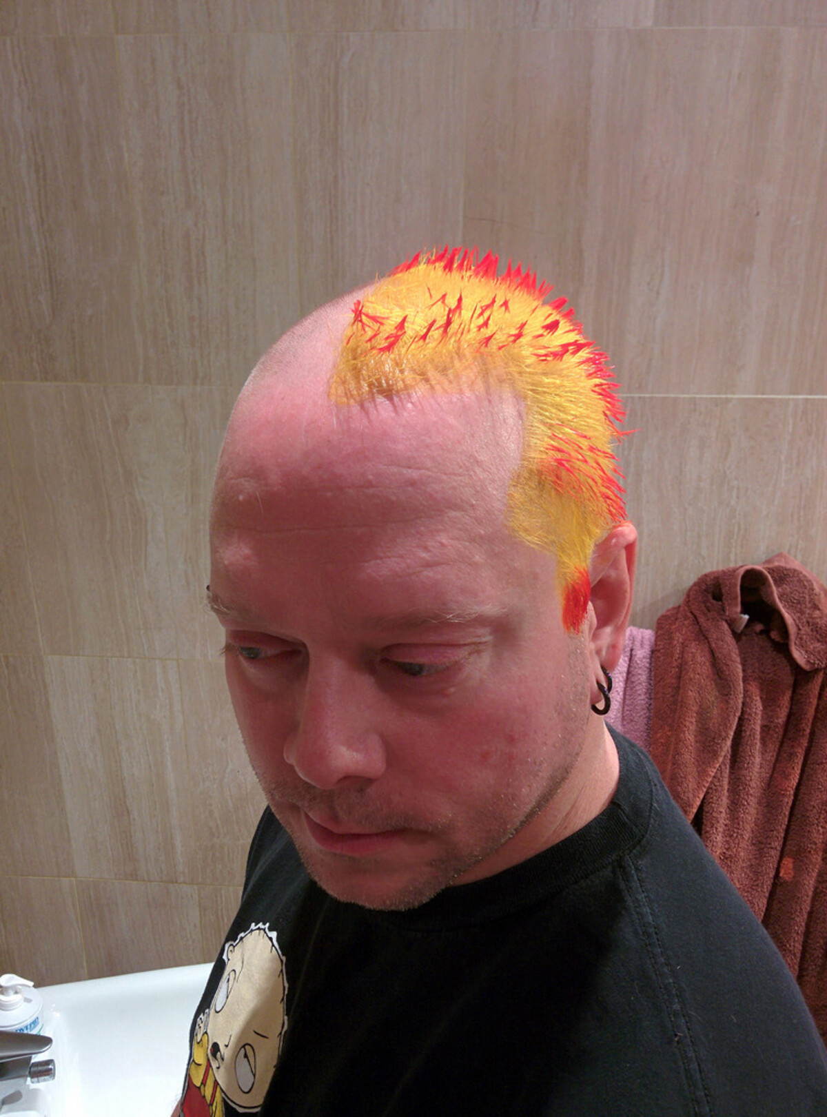 Rob in the bathroom looking down, a bright yellow-orange mohawk with red-tipped spikes standing upright across his head. Sides shaved. Black ear gauge, cartoon t-shirt. Bathroom tiles and a brown robe behind him.