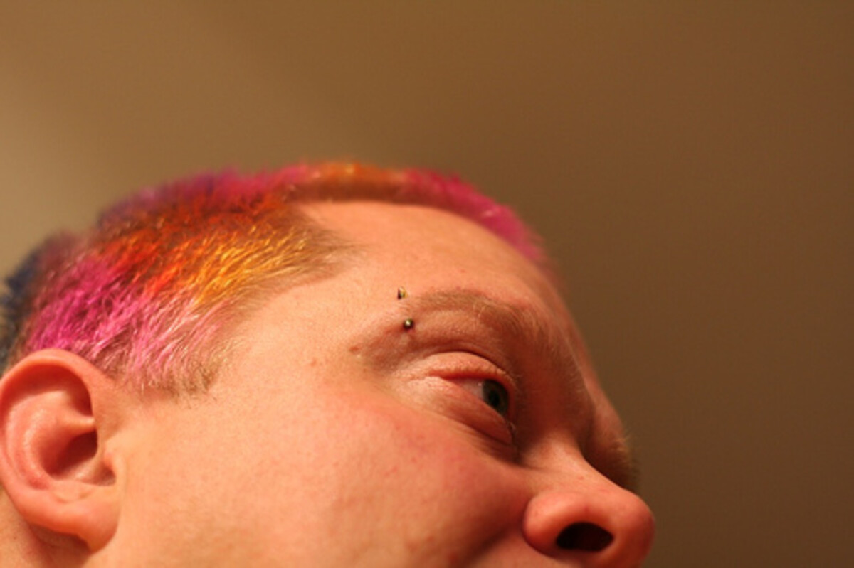 Close-up of Rob's face from above, showing a gradient of hot pink, orange, and blue-purple hair in a short buzz. Bridge piercing visible.