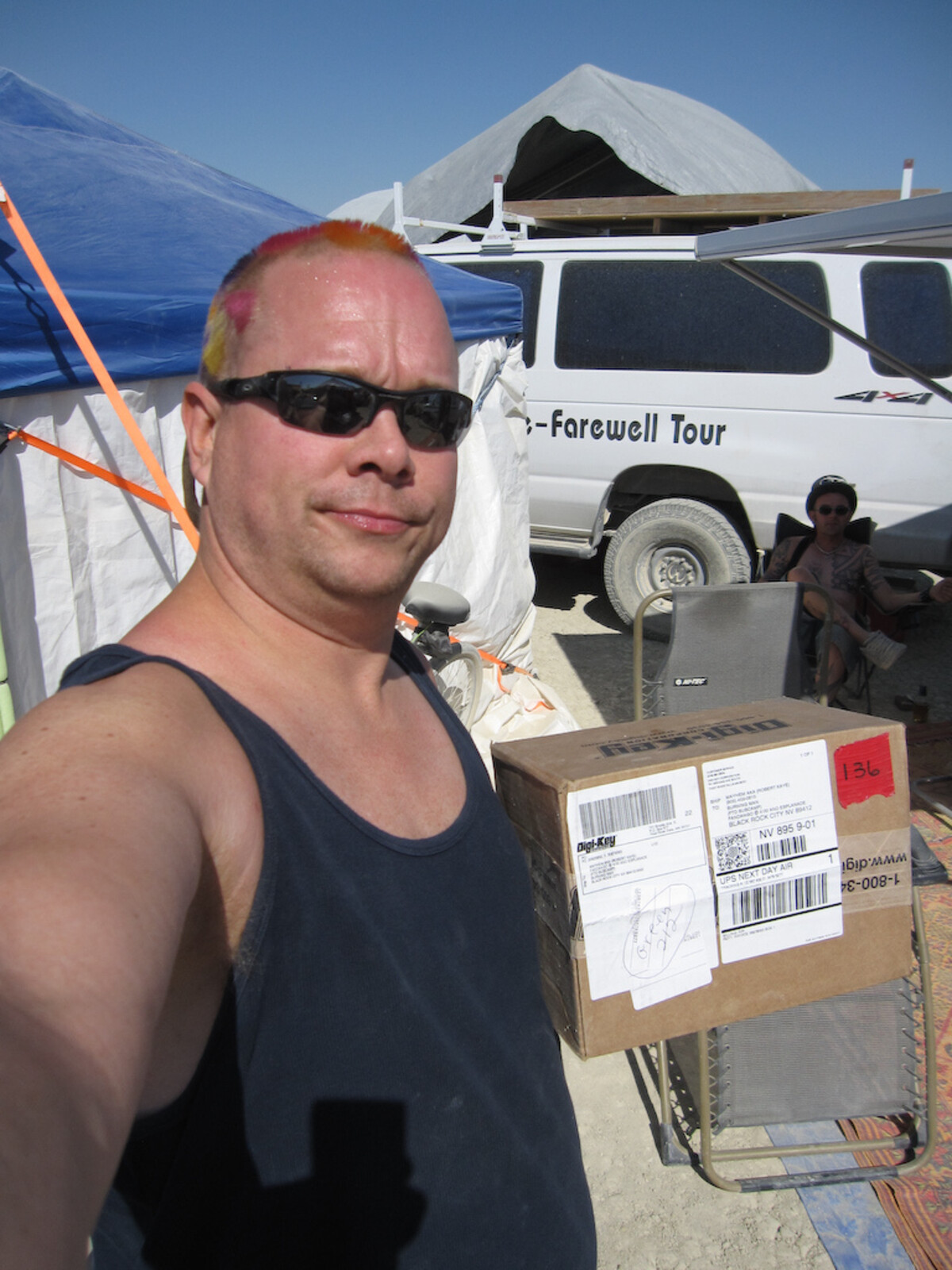 Rob at Burning Man in a tank top and sunglasses, faded pink-orange hair tufts on his crown, standing near a Digi-Key box and a van reading 'Farewell Tour.'