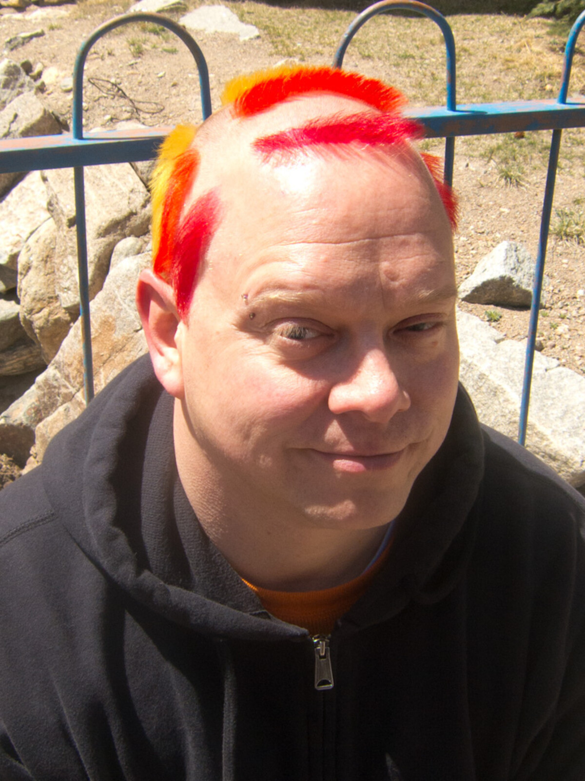 Rob sitting outdoors against a blue metal railing, short buzzed hair in bold red and yellow-gold stripes, wearing a dark hoodie. Bridge piercing visible, hint of a smile.