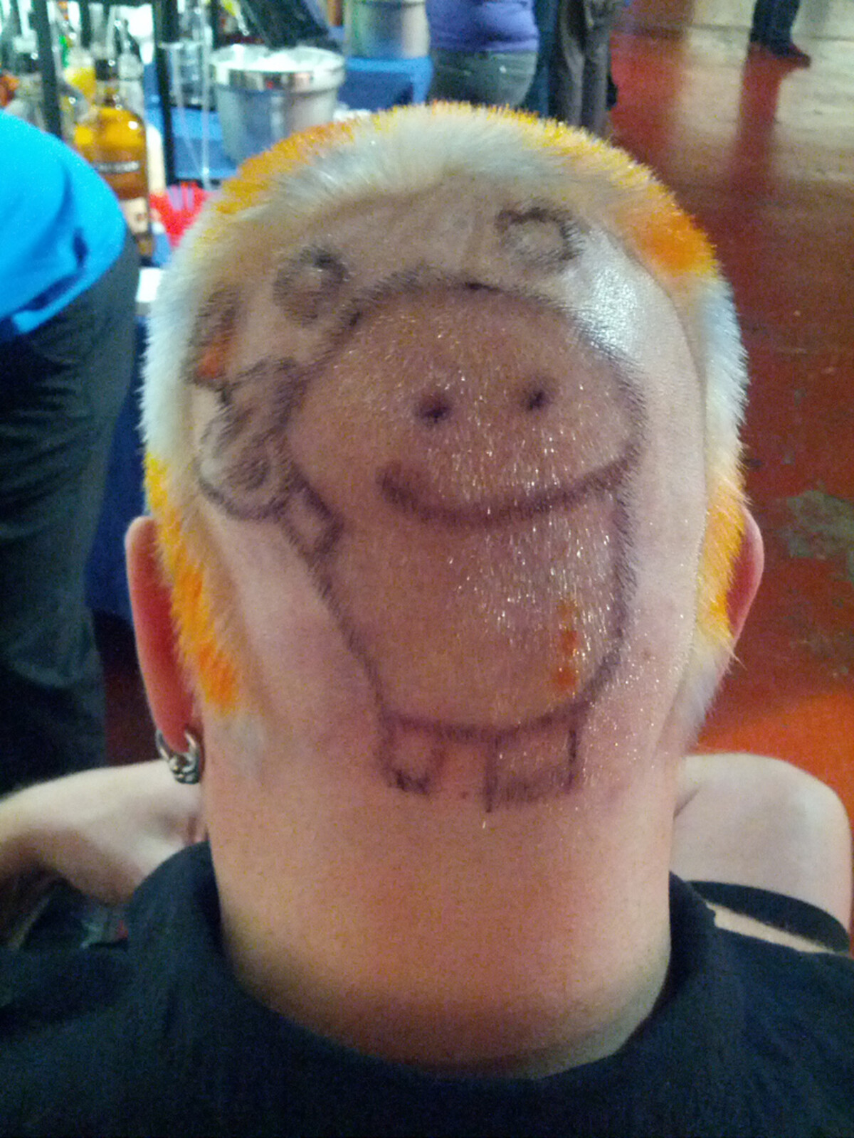 Back of Rob's head with a cartoon robot character shaved and drawn onto the scalp &mdash; boxy body, smiling face, antenna &mdash; framed by orange-yellow dyed hair at the edges. Bar setting in background.