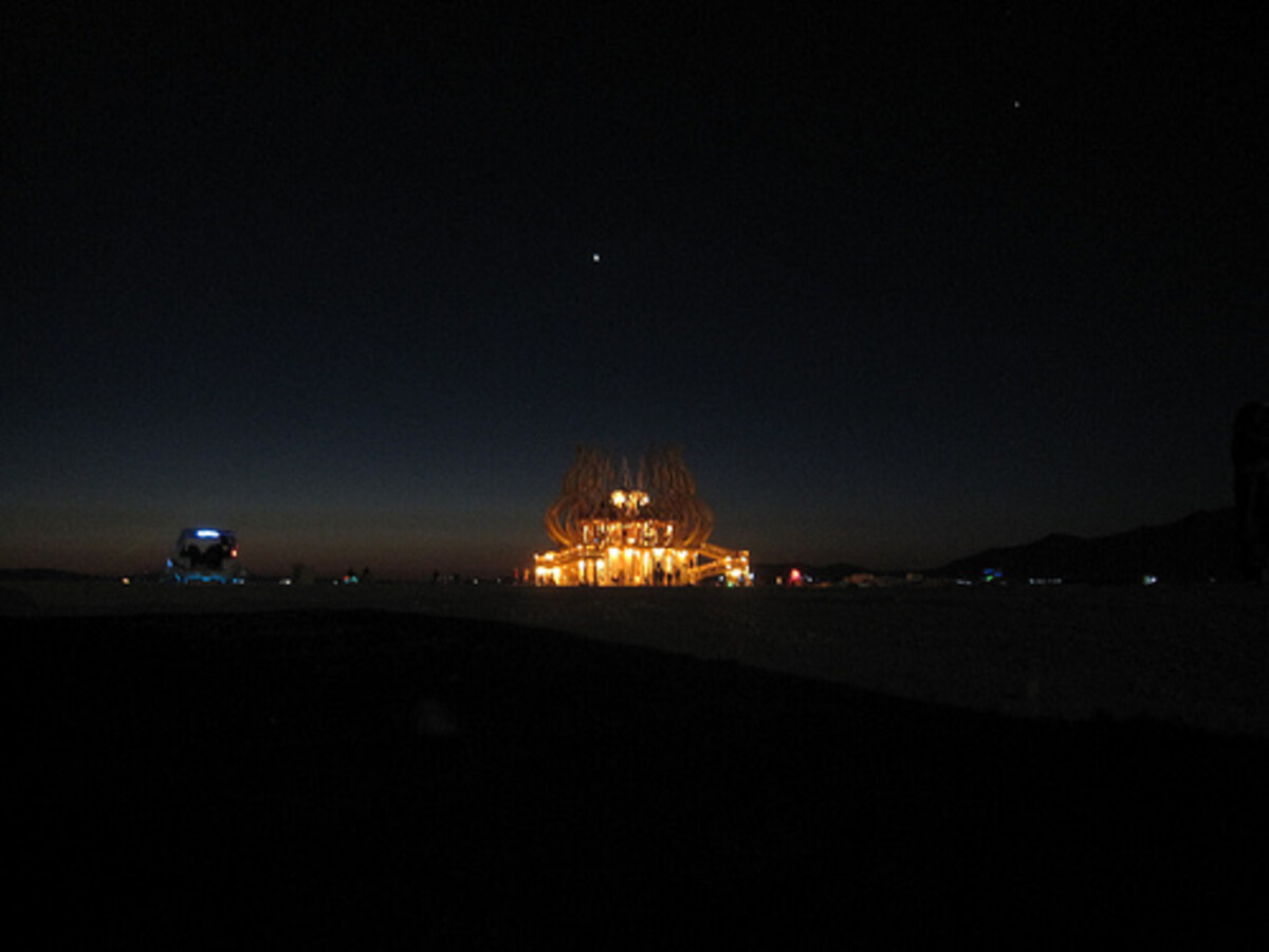 A Burning Man memorial temple glowing golden against a dark night sky on the playa, stars visible overhead, a distant lit art car to the left.