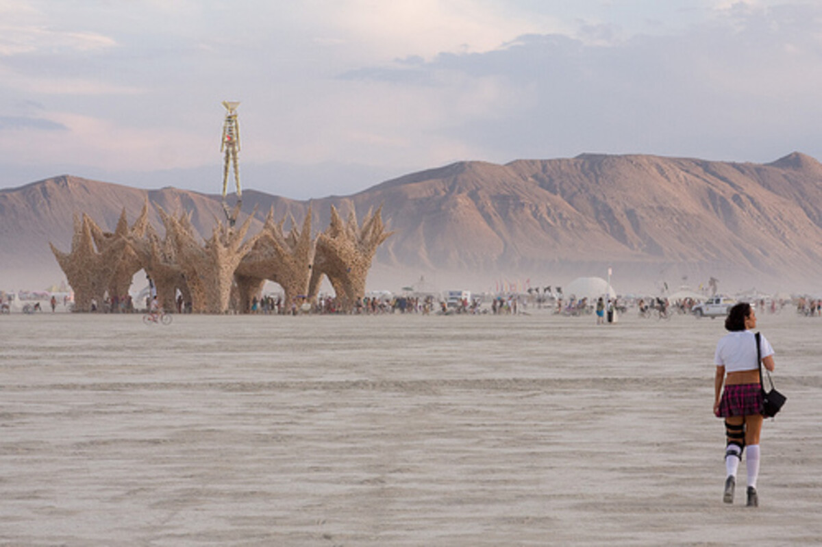 A person in a white t-shirt, plaid skirt, and knee-high socks walking alone across the open playa toward the 2009 Burning Man temple, a towering organic lattice structure, with the Man figure on his tower behind it and desert mountains on the horizon.