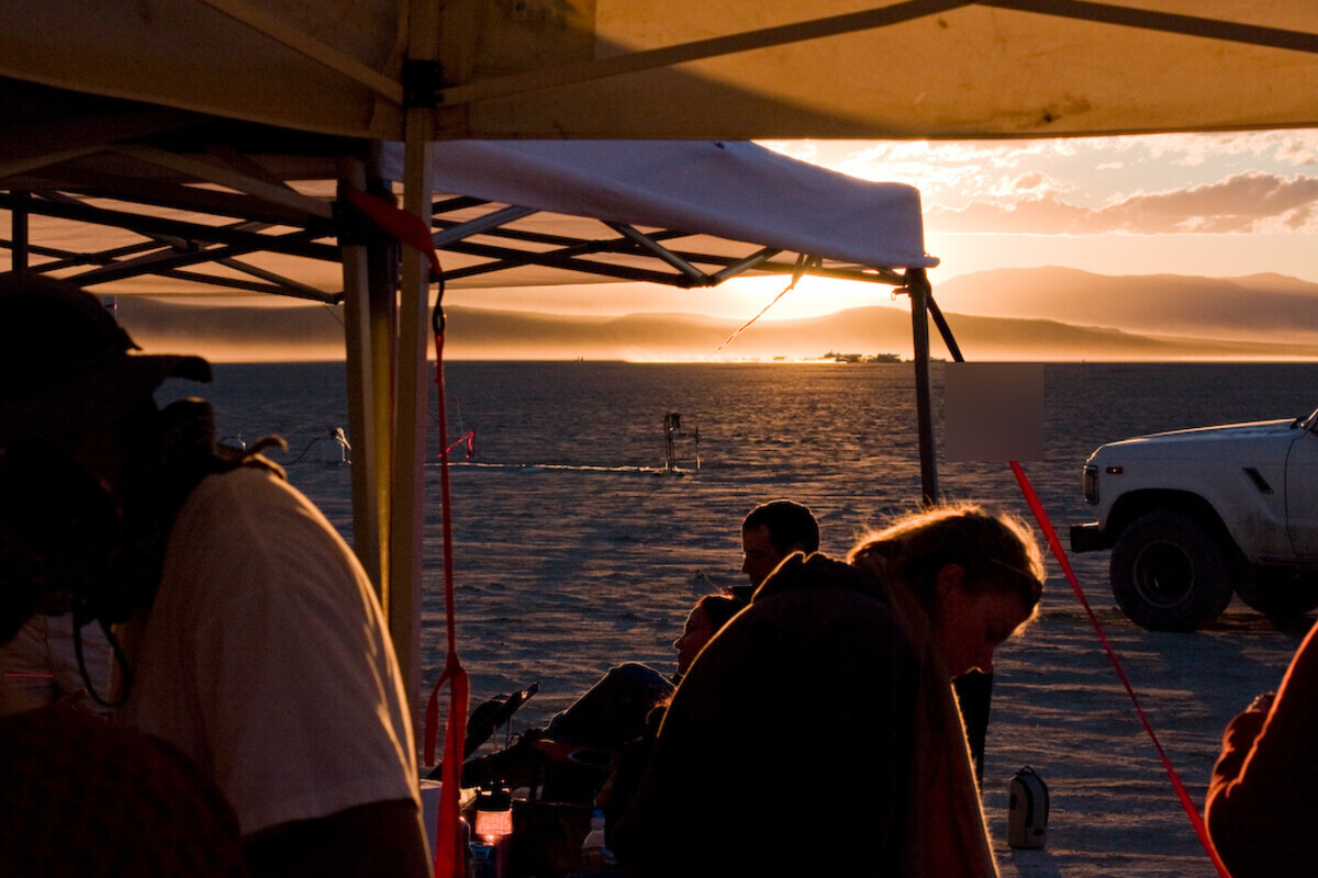 Sunset over the desert playa seen from under a shade canopy at the 4th of Juplaya burn, people silhouetted against golden light, mountains and low dust haze on the horizon, a pickup truck at the edge of camp. Face blurred.