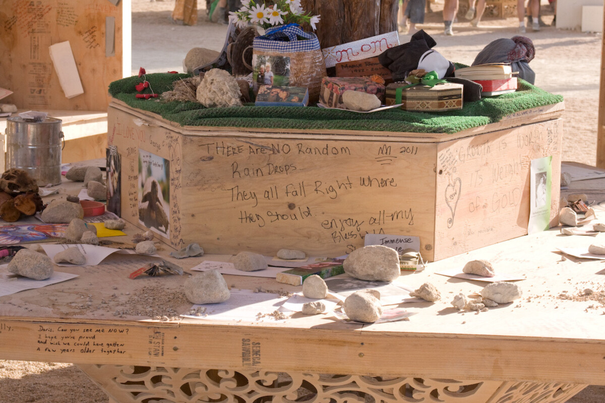 A wooden shrine at the 2011 Burning Man temple covered with rocks, wildflowers, photographs, and small keepsakes on green felt, with handwritten messages including 'There are no random raindrops — they all fall right where they should.'