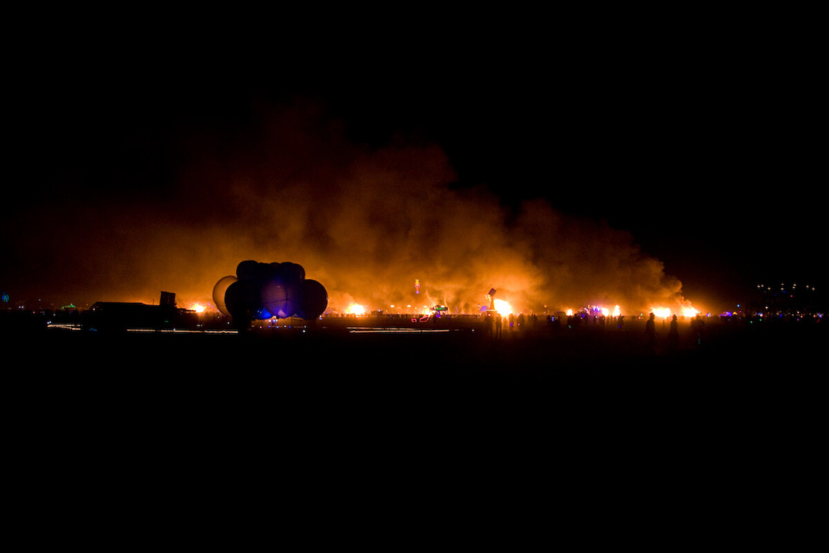 Wide panoramic night shot at Burning Man 2011: a line of fire along the entire horizon, art installations silhouetted against the glow including a cloud sculpture lit purple, thick smoke and orange light filling the sky, tiny scattered figures on the playa.