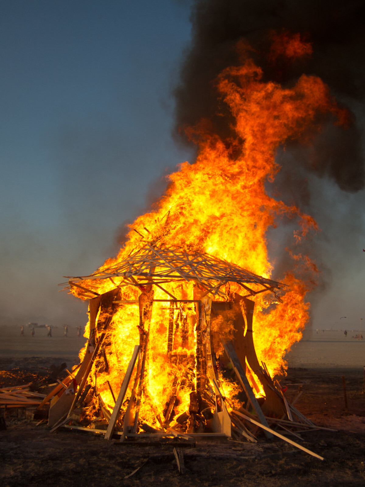 A wooden memorial temple fully engulfed in towering yellow-orange flames and black smoke, the lattice frame still visible as it collapses inward, small figures on the playa in the background.
