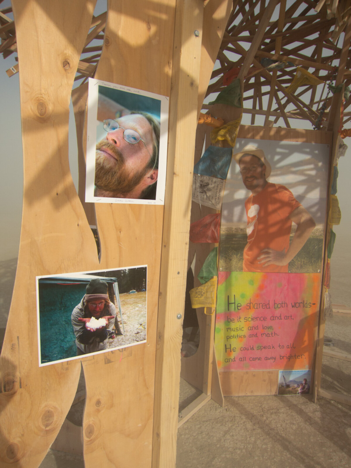 Close-up of wooden temple panels with three photographs of a bearded man with glasses pinned to the wood, a colorful handpainted poem below reading 'He shared both worlds,' and prayer flags overhead.