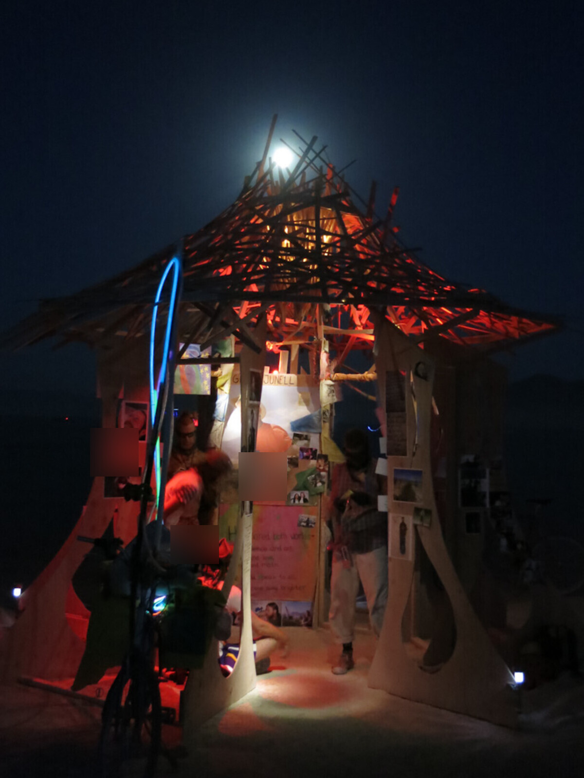 Greg's memorial temple at night on the playa, the full moon rising through the open lattice roof, visitors gathered inside among photographs and tributes, 'JUNELL' visible on a panel, a bicycle and a glowing blue LED hoop at the entrance. Faces blurred.