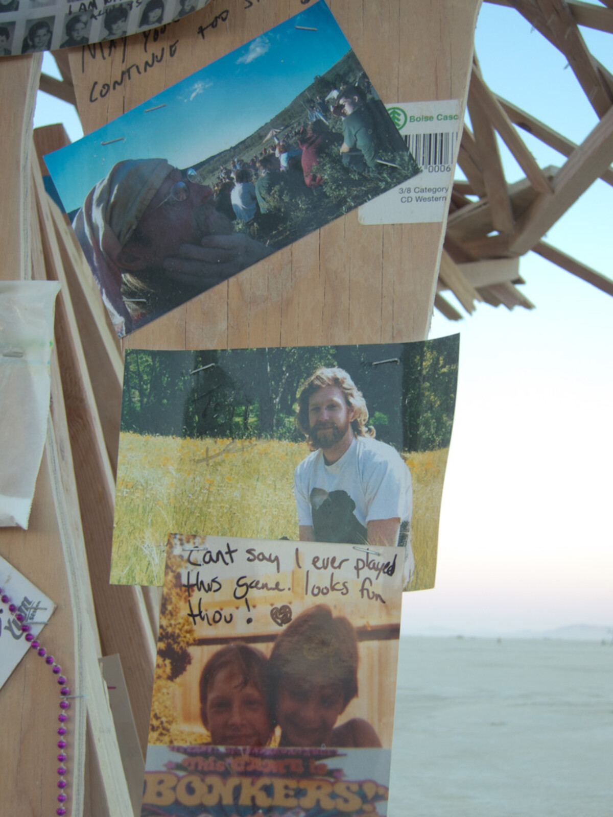 Close-up of photographs and handwritten notes pinned to a wooden temple panel — a bearded man standing in a golden field, a group photo, and a handwritten note with a heart, the playa visible through the lattice behind.