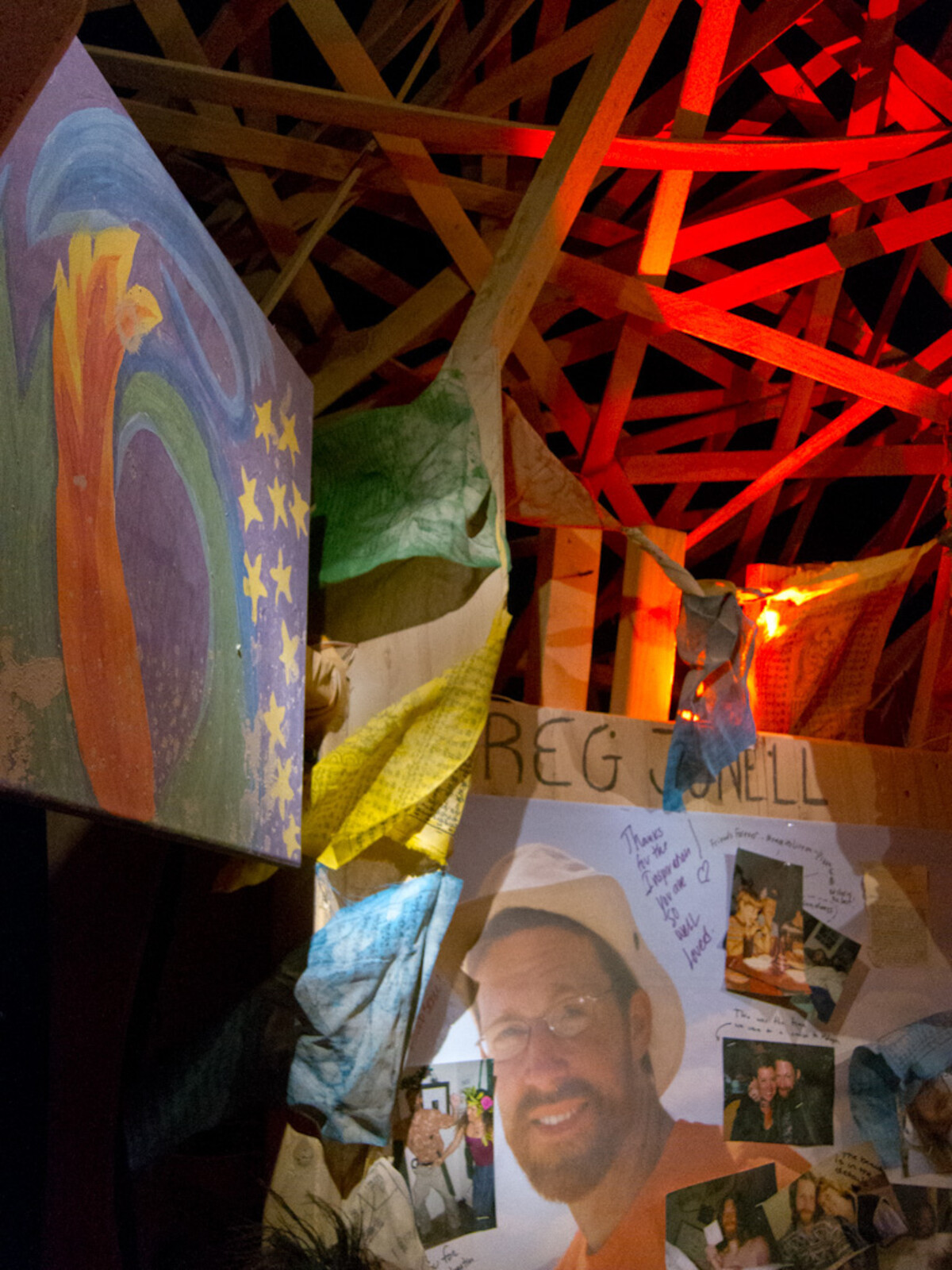 Interior of a wooden lattice temple at night lit by deep red light, 'GREG JUNELL' written in large letters on a memorial board, photographs of a smiling man below, a painted rainbow artwork to the left, cloth banners hanging overhead.