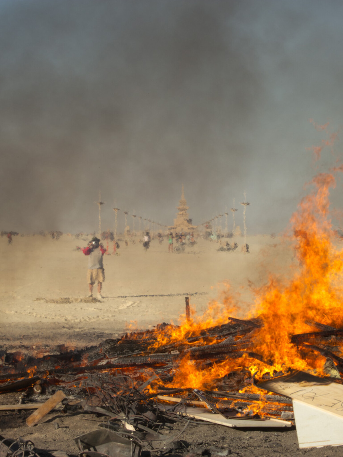 Burning temple remains in the foreground with bright orange flames, a lone figure standing in the smoky middle distance photographing, the main Burning Man temple barely visible through heat shimmer and thick smoke behind.