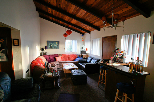 Living room with exposed wood beams, leather couches in blue and red, red Chinese lanterns, a bar area with bottles, and warm eclectic decor.