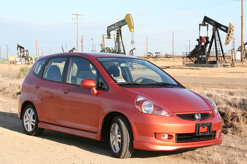Orange Honda Fit parked in a dusty field with oil pumpjacks visible in the background against a Central California landscape.