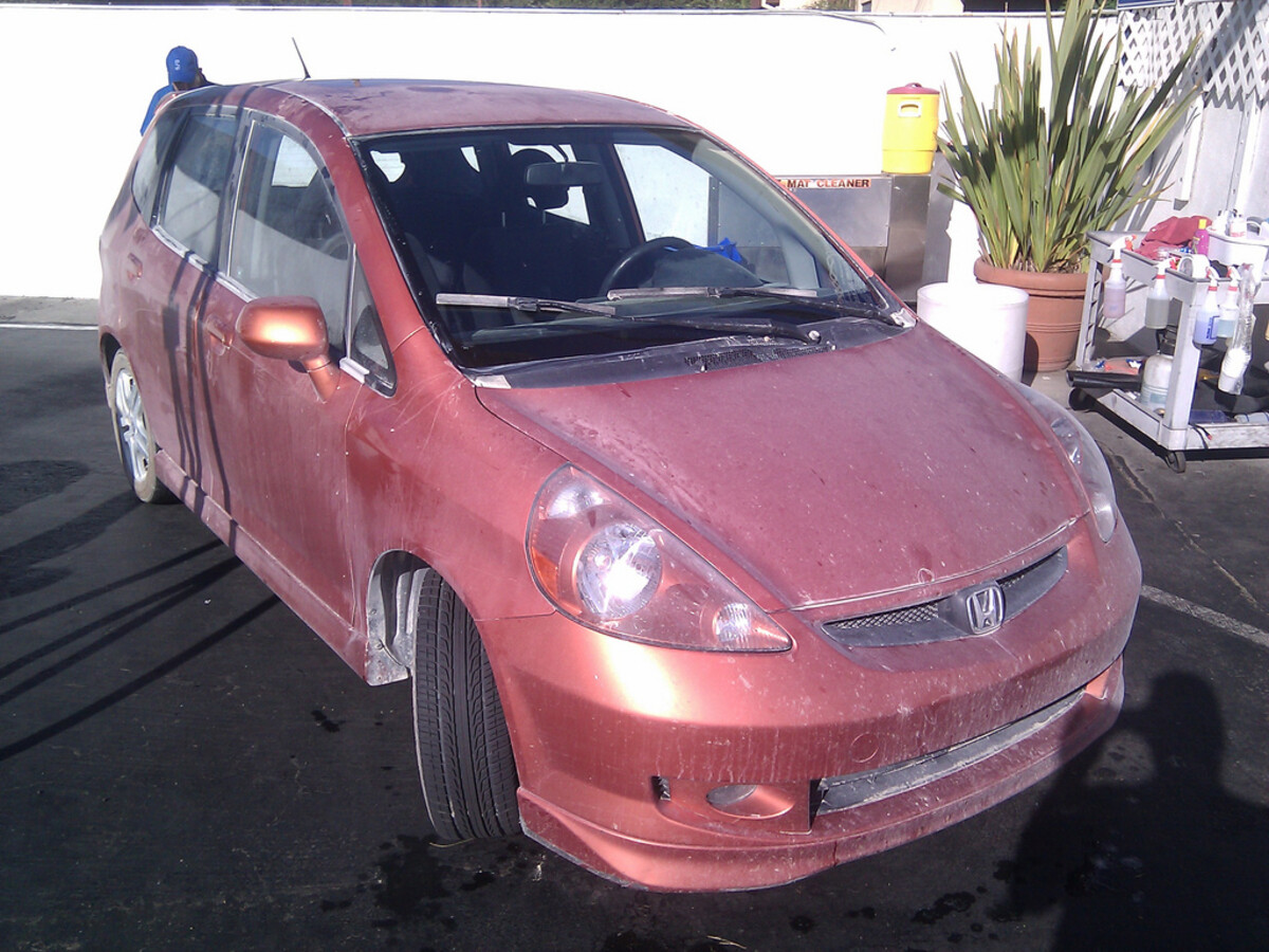 Orange Honda Fit covered in thick playa dust at a car wash, headlights barely visible through the grime.