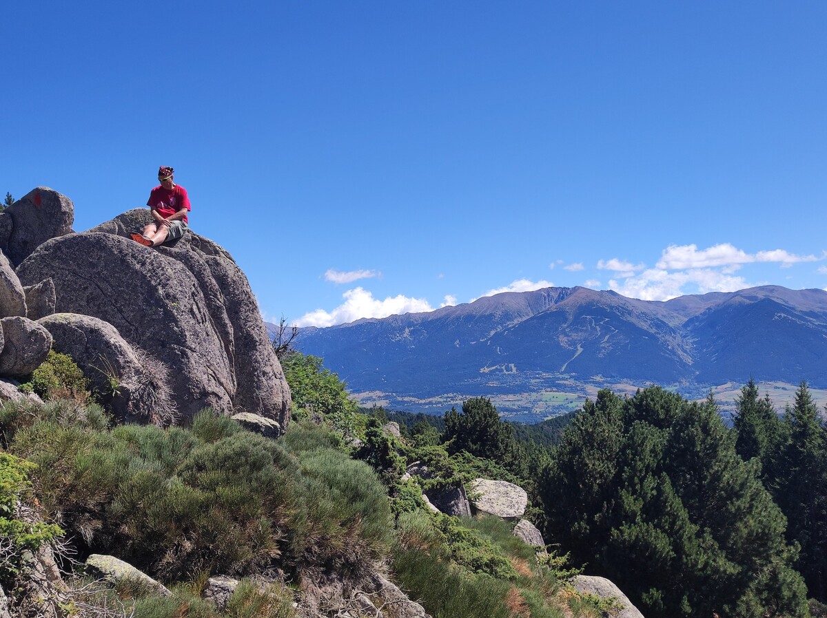 Rob in a red t-shirt and cap sitting cross-legged on a large boulder high on a mountainside, panoramic view of a green valley and the Pyrenees range stretching across the background under a deep blue sky with scattered clouds. Font-Romeu, France.