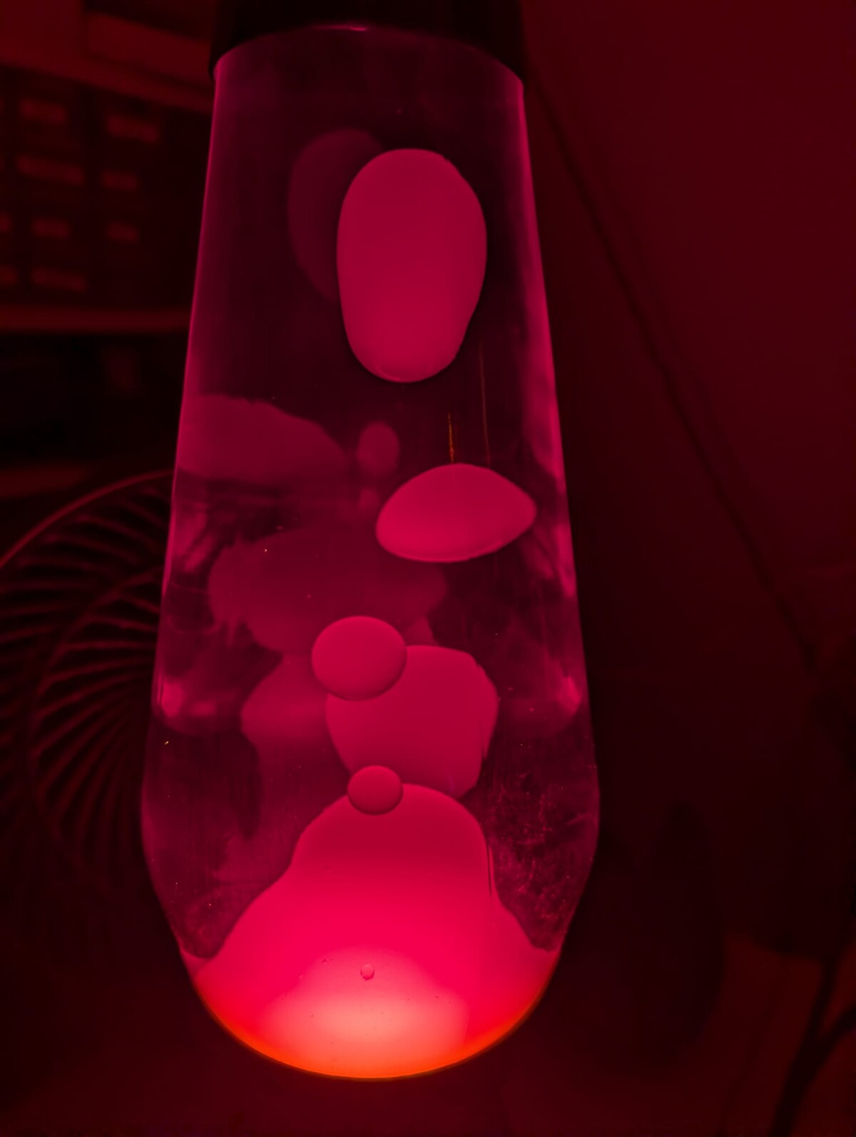 Close-up of a lava lamp glowing hot pink and red in a dark room, large wax blobs floating and merging, casting a deep magenta light across the surrounding darkness.