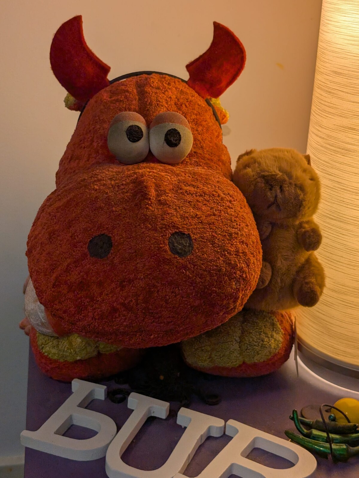 Large red stuffed hippo with felt horns and googly eyes sits on a shelf beside a small brown plush capybara, a lamp glowing behind them, decorative letters spelling BUR visible below.