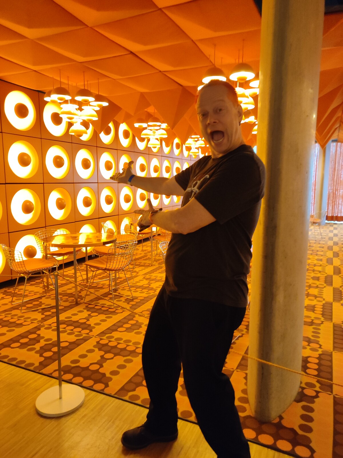 Rob mid-exclamation, arms spread wide, mouth open in exaggerated delight, standing inside the Spiegel Kantine &mdash; a retro-futuristic 1960s canteen with bright orange padded ceiling, a grid of circular backlit speaker-like panels on the wall, wire chairs, and an orange polka-dot floor. Museum f&uuml;r Kunst und Gewerbe, Hamburg.