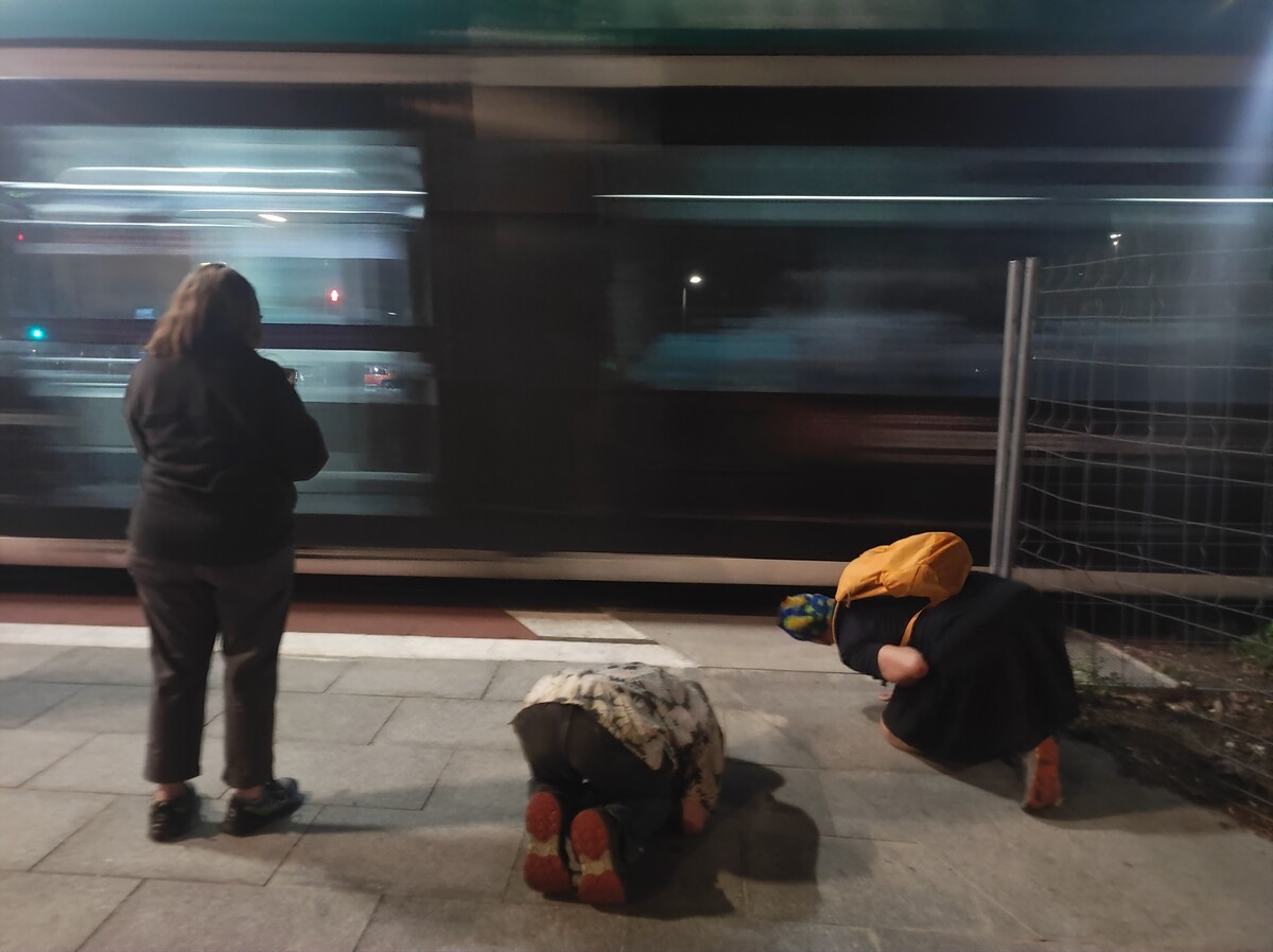 Night scene at a Barcelona tram stop: two people crouch on the platform peering under the tracks of a passing tram, motion-blurred green and white, while a woman stands watching to the left. Yellow backpack visible on one of the crouching figures.