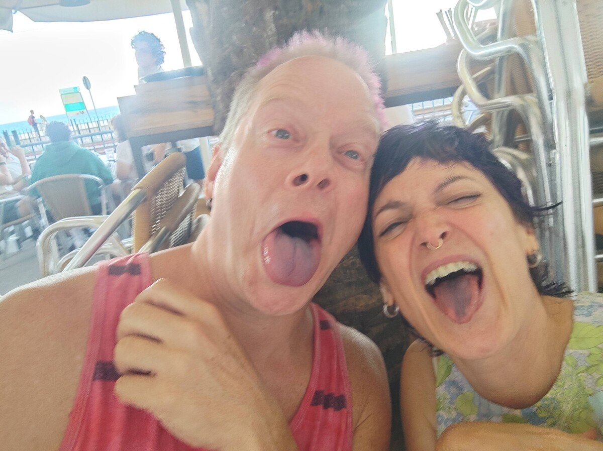 Close-up selfie of Rob and Gisela at a beachside restaurant, both sticking out purple-stained tongues and laughing wide, Rob in a pink tank top with faint pink hair, Gisela with short dark hair and a nose ring, outdoor seating and other diners behind them.