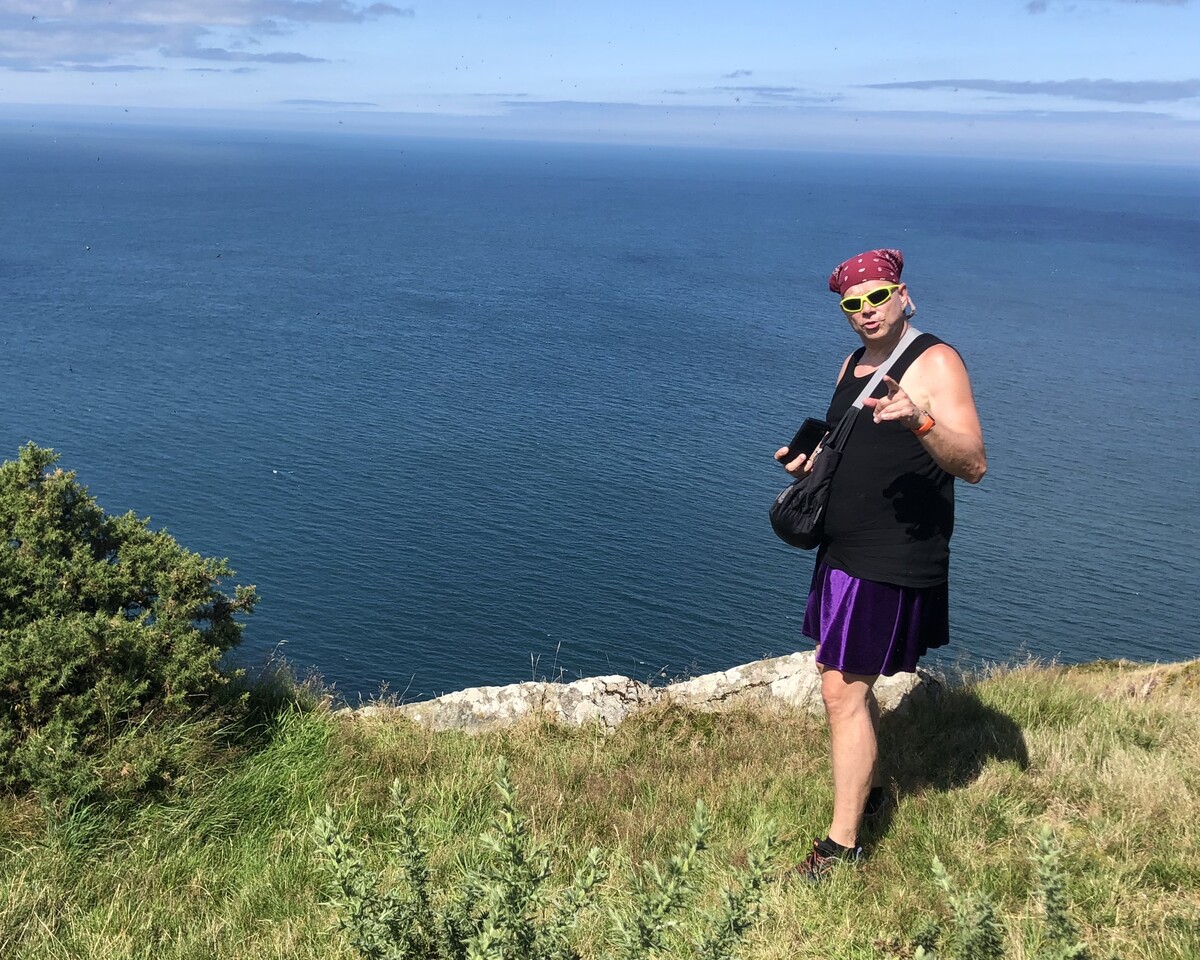 Rob on a cliff edge overlooking deep blue sea, wearing a purple kilt, pink bandana, and neon yellow-green sunglasses, gesturing outward with one hand, green cliff-top vegetation and blue sky around him.