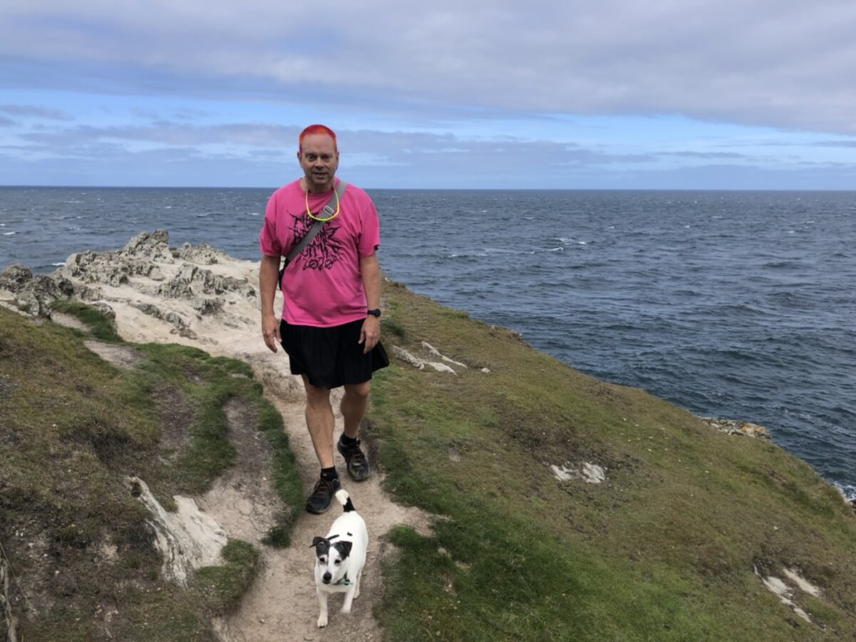 Rob in a hot pink graphic t-shirt and black shorts standing on a narrow coastal path, orange-red hair visible, yellow-green sunglasses around his neck, a Jack Russell terrier ahead on the trail, rocky outcrop and grey-blue sea behind him.