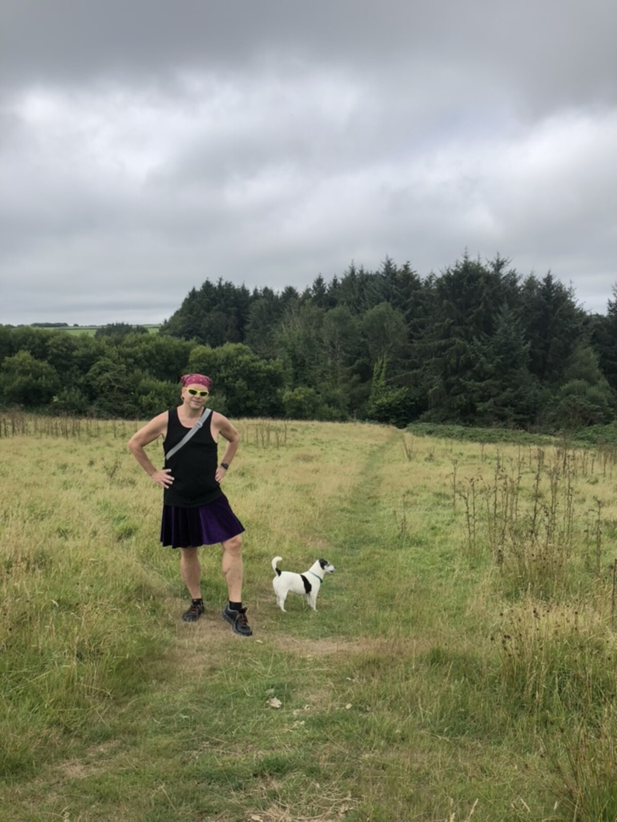 Rob standing hands on hips on an English countryside footpath in a purple kilt, black tank top, pink bandana, and neon yellow-green sunglasses, a Jack Russell terrier beside him, rolling green meadow and overcast sky behind.