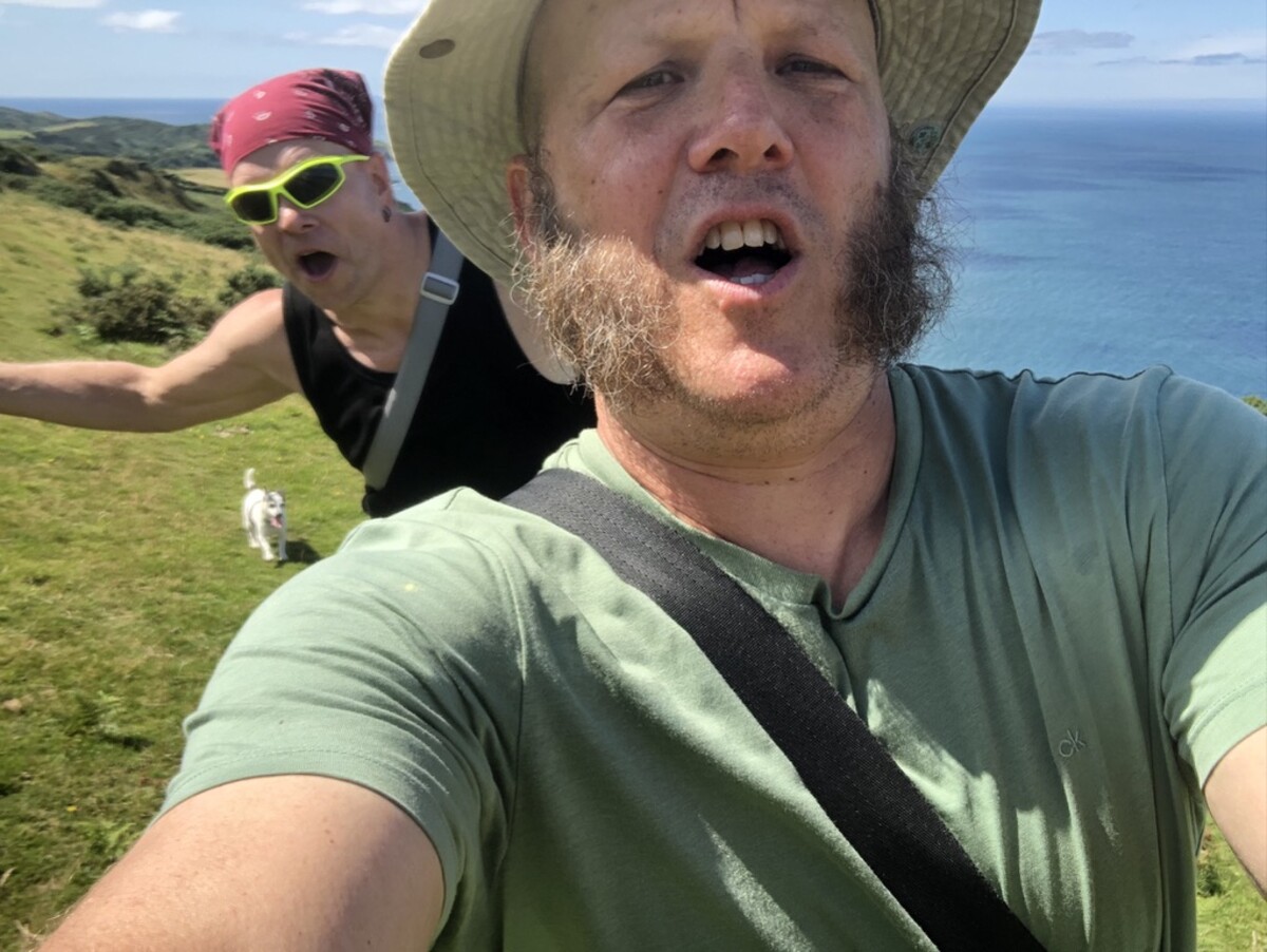 Selfie of Ian in a bucket hat and Rob in a pink bandana and yellow-green sunglasses, both with mouths wide open in exaggerated yells, a Jack Russell terrier on the green hillside between them, turquoise sea and coastal hills behind.