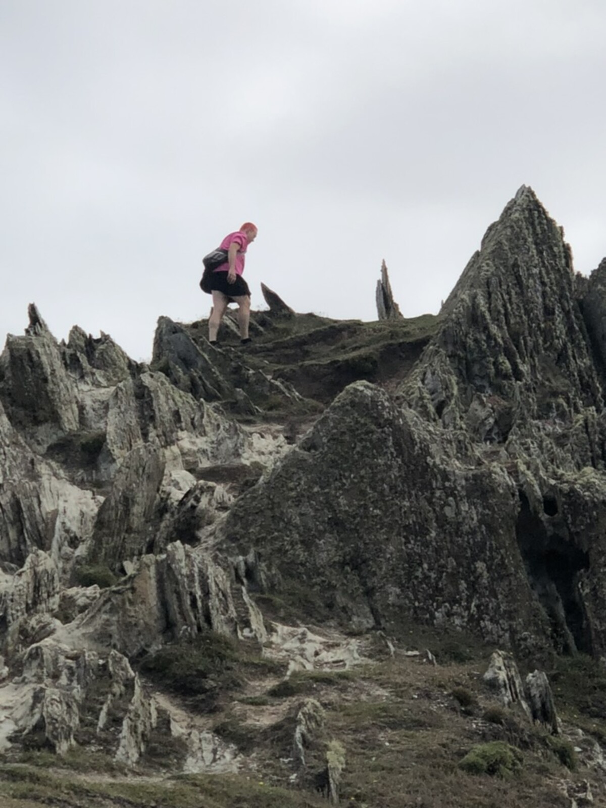 Small figure in a pink shirt walking away along a narrow ridge among dramatic jagged slate rock formations, a single bright dot of color against ancient grey stone and overcast sky. England, August 2024.