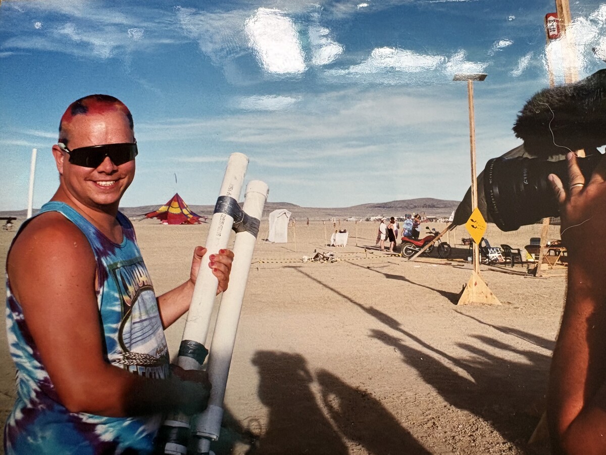Young Rob with pink-red hair and a tie-dye tank top, grinning and holding two white PVC pipe assemblies on the Burning Man playa, camp structures and a dramatic desert sky behind him. Scanned print with visible creasing.