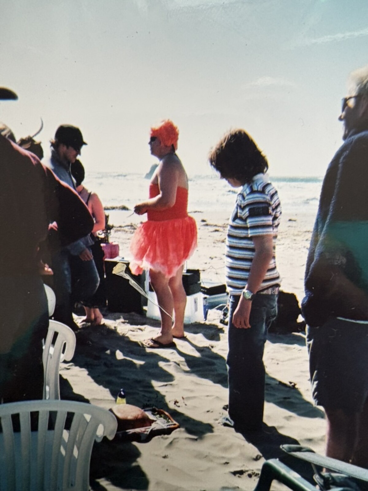 Rob standing barefoot on a beach among wedding guests in a bright orange tutu and matching tube top, vivid orange hair, sound equipment cases on the sand beside him, ocean and hazy sky behind. Cayucos, California.