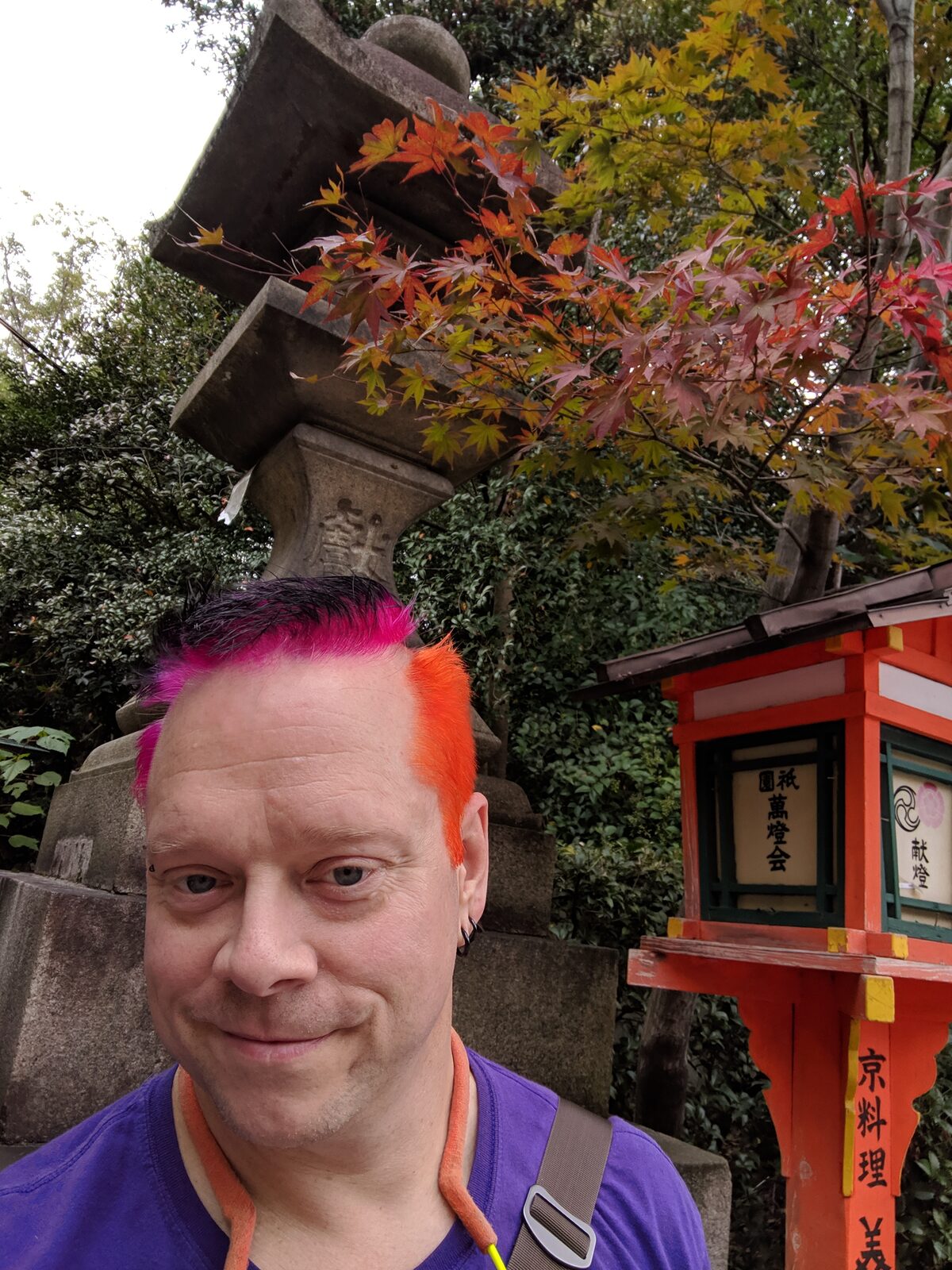 Rob at a Japanese shrine in autumn, pink and orange split mohawk standing upright, wearing a purple t-shirt. Stone lantern, orange Shinto lamp, and red maple leaves frame him above.