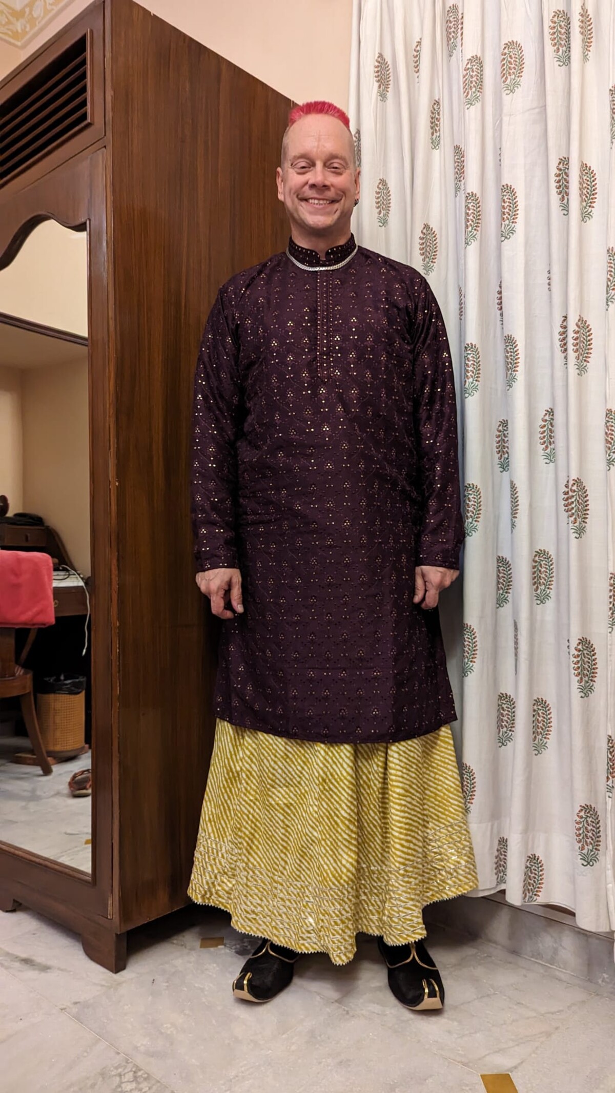 Rob standing in a Jaipur bedroom wearing a purple kurta with gold embroidery and a golden sharara, black juttis on his feet, pink mohawk on top, grinning with eyes closed.