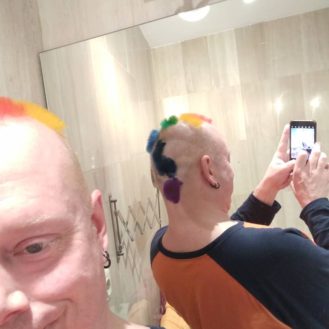 Rob in a bathroom mirror selfie showing both front and back of his shaved head: red-orange-yellow patches at the front, green, blue, and purple patches at the back. Wearing a navy and orange raglan shirt, holding phone to capture the mirror view.