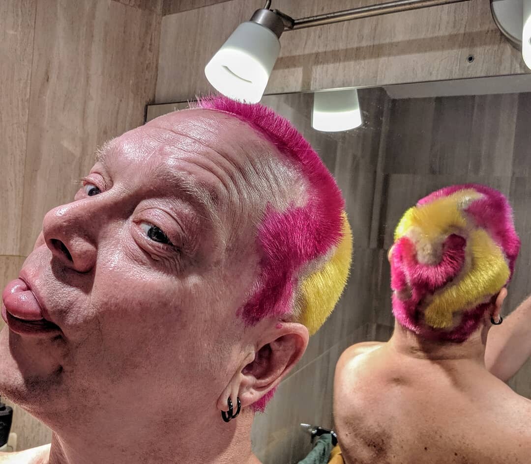 Rob in a bathroom selfie sticking his tongue out, hot pink and yellow concentric spiral pattern dyed onto his buzzed head. The bathroom mirror behind shows the back of the design swirling from the crown. Black hoop earrings.
