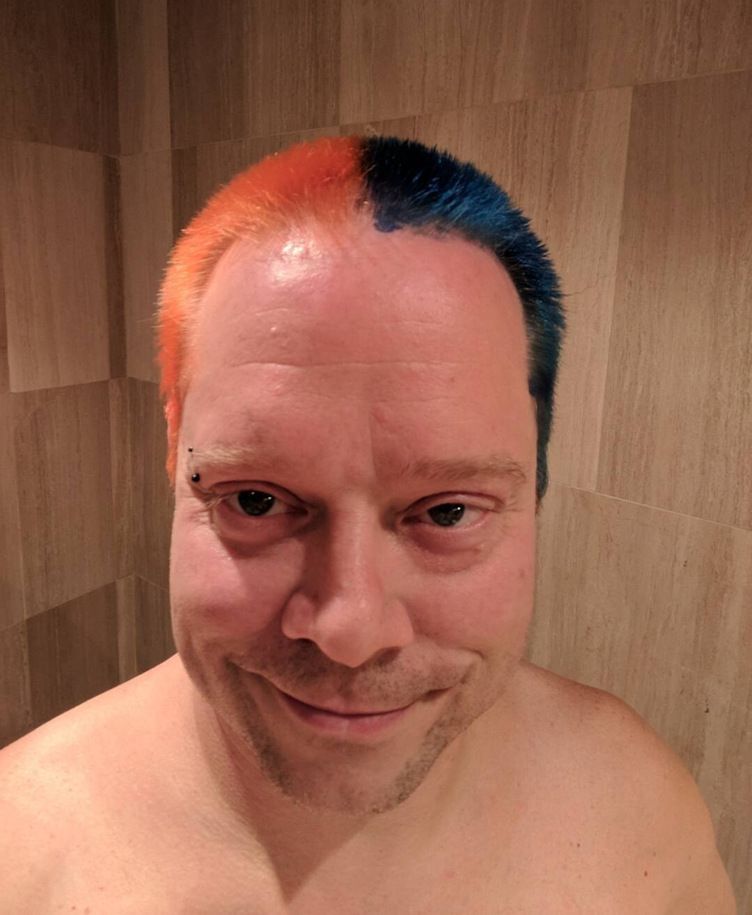 Rob in a bathroom selfie, hair split cleanly down the middle: bright orange on the left side, deep teal-blue on the right. Bridge piercing visible, slight smirk.