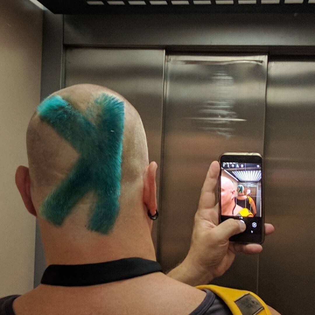 Back of Rob's shaved head with a bold teal X dyed across the scalp, photographing himself in an elevator with his phone. The phone screen shows the same X from the front. Black earring visible.