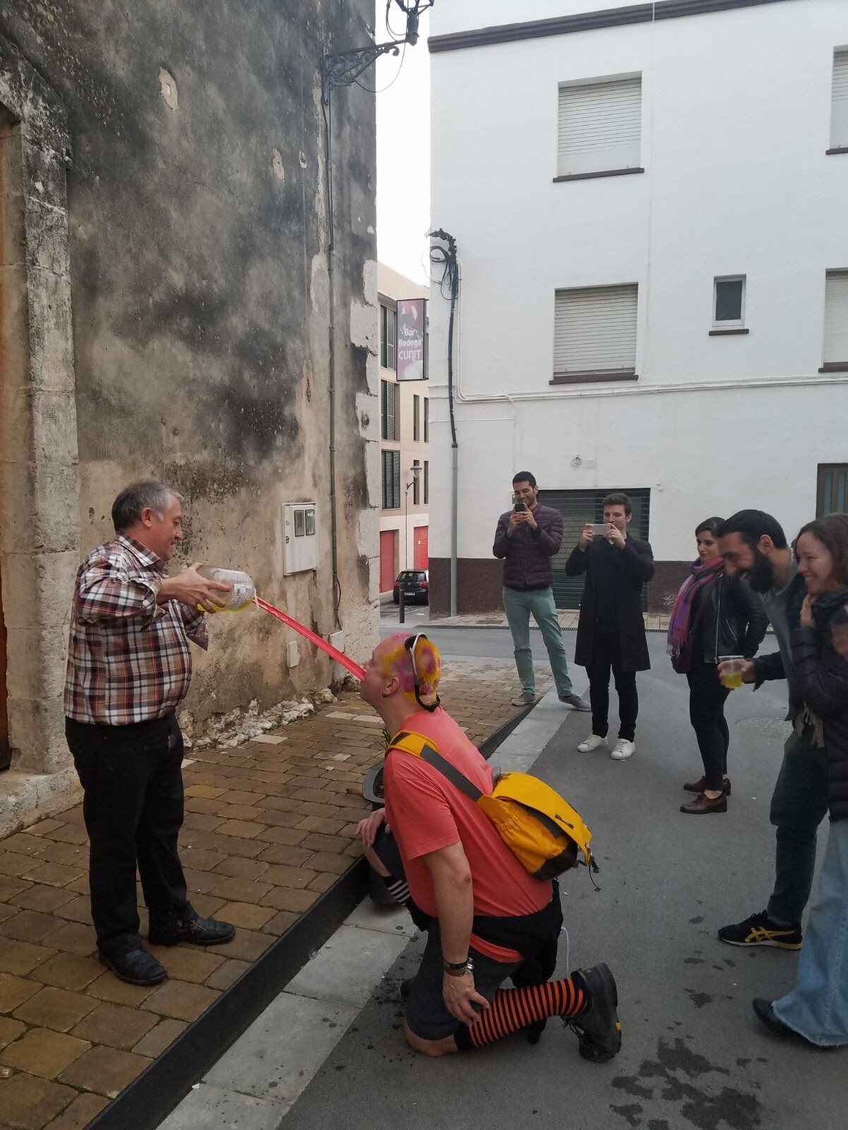 Rob kneeling on a Catalan village street, head tilted back, drinking wine poured through a red funnel by a man in a plaid shirt. Pink and yellow hair already dyed. Friends stand behind filming and laughing. Orange-and-black stripy socks.