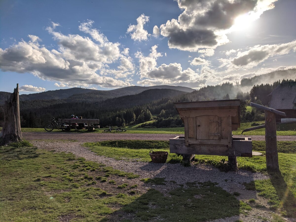 Wide landscape of a Black Forest farmstead: a wooden trough structure (the 'beer manger') in the foreground, rolling green hills, farm buildings, and dramatic cumulus clouds backlit by the sun.