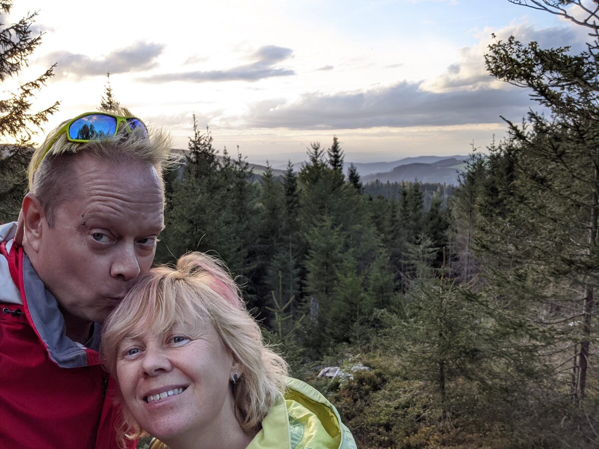 Rob with platinum hair and blue reflective sunglasses and Kirsten in a lime-green windbreaker, leaning together for a selfie on a Black Forest hilltop with dense conifer forest stretching to the horizon.