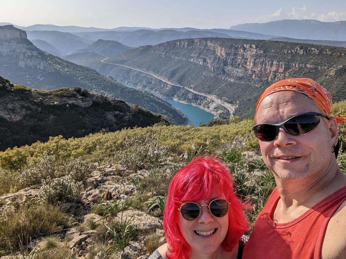 Selfie of Rob in an orange bandana and Kirsten with vivid red-magenta hair at a high viewpoint over a deep limestone canyon with layered mountain ridges receding into haze and a turquoise river visible far below.