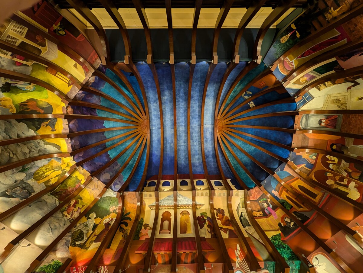 Ornate painted ceiling with a central blue-and-gold sunburst radiating pattern, surrounded by colorful narrative murals depicting Indian cultural scenes of agriculture, nature, and daily life.