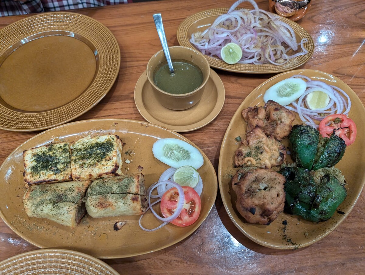 Overhead flat-lay of four clay plates of Indian street food including grilled sandwiches, tikka with sliced onion and lemon, stuffed peppers, and a cup of green chutney on a wooden table.