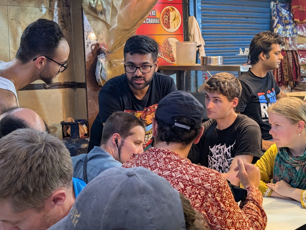 Crowded scene at a Delhi street food stall with approximately nine people packed around a table, Hindi signage visible. Rob partially visible by his pink hair and rust-patterned kurta, turned away from the camera.