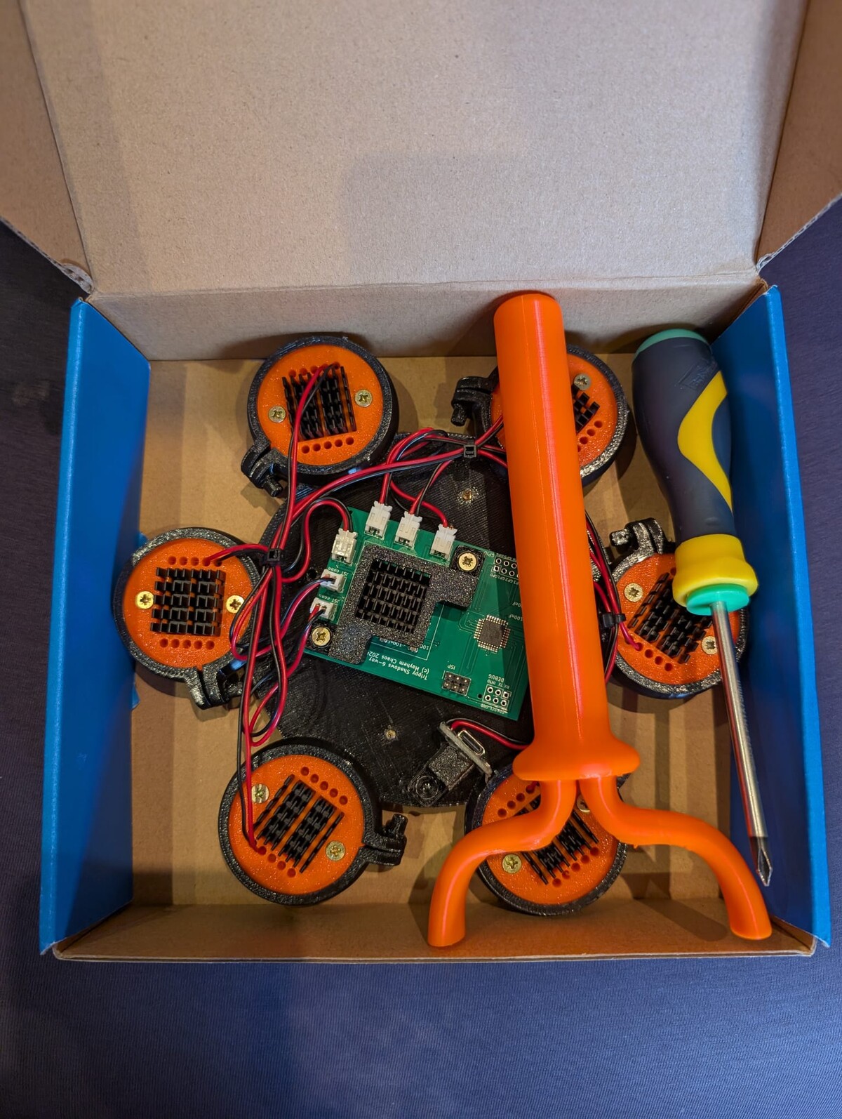 Top-down view into a cardboard box containing a custom-built juggling device with multiple orange speaker-like elements, a green circuit board with wiring, and orange 3D-printed parts.
