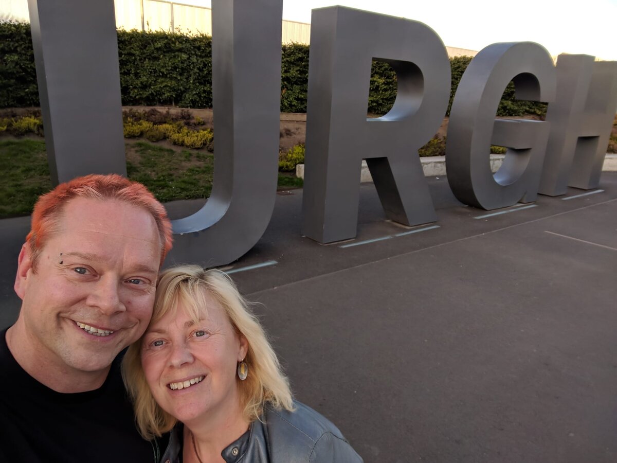 Rob with orange-red hair and Kirsten with blonde hair, cheek to cheek and grinning broadly, in front of large 3D metal letters spelling EDINBURGH.
