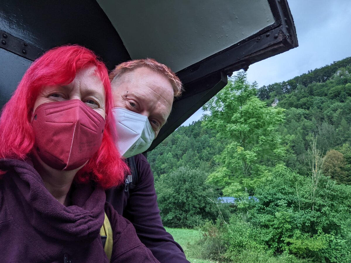 Rob and Kirsten in FFP2 masks taking a selfie from inside a heritage steam train carriage, crinkled smiling eyes visible above the masks, green forested hills of Franconian Switzerland through the window behind them.